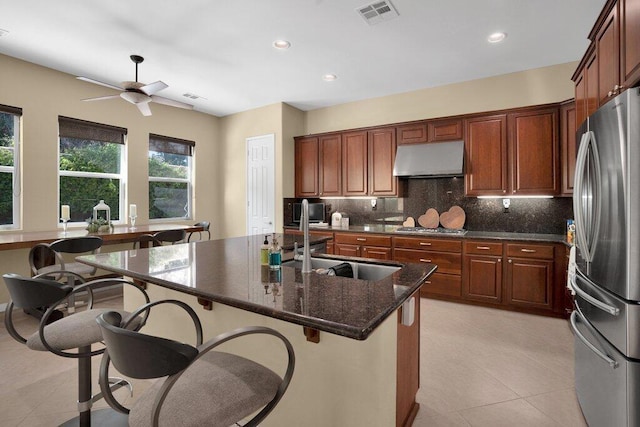 kitchen with ceiling fan, backsplash, dark stone countertops, a center island with sink, and appliances with stainless steel finishes