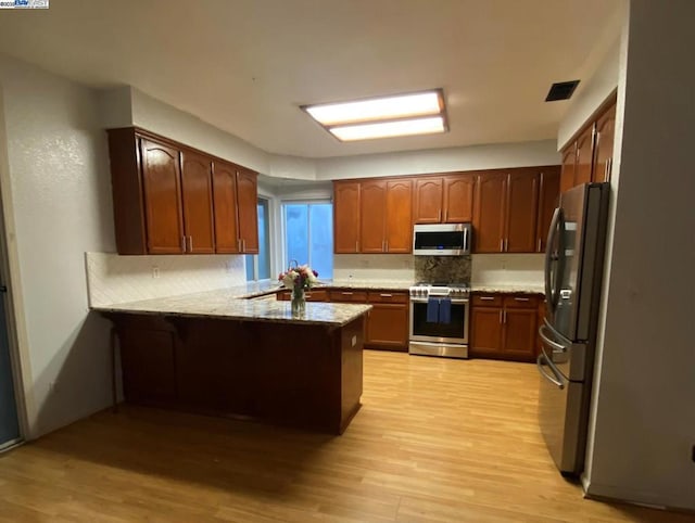 kitchen with kitchen peninsula, light stone countertops, light wood-type flooring, tasteful backsplash, and stainless steel appliances