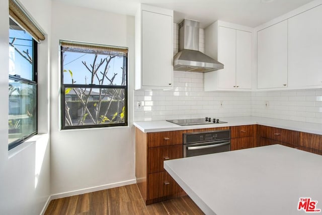 kitchen featuring black electric stovetop, white cabinets, oven, wall chimney range hood, and tasteful backsplash