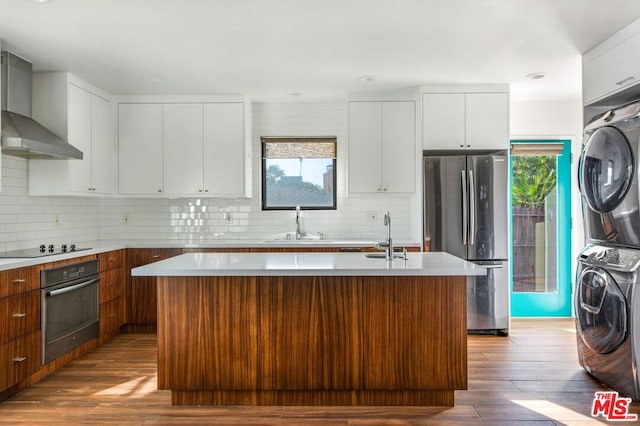 kitchen featuring a center island with sink, wall chimney range hood, oven, stainless steel refrigerator, and stacked washer and clothes dryer