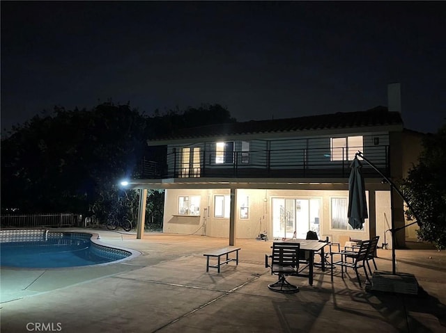 back of house at night with an outdoor pool, stucco siding, a patio, and a balcony