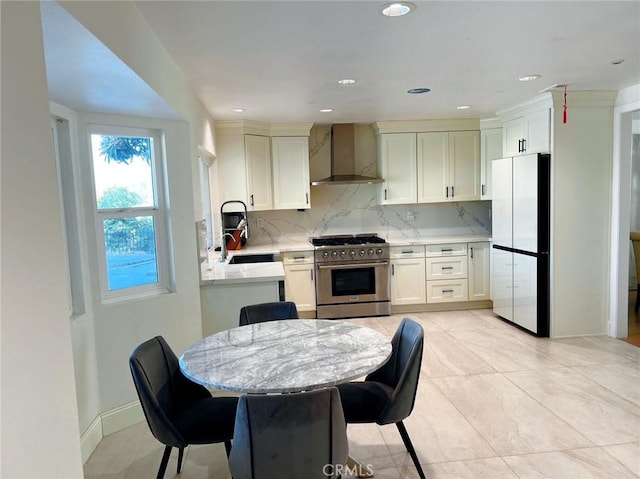 kitchen with freestanding refrigerator, wall chimney exhaust hood, high end stainless steel range, light stone countertops, and backsplash