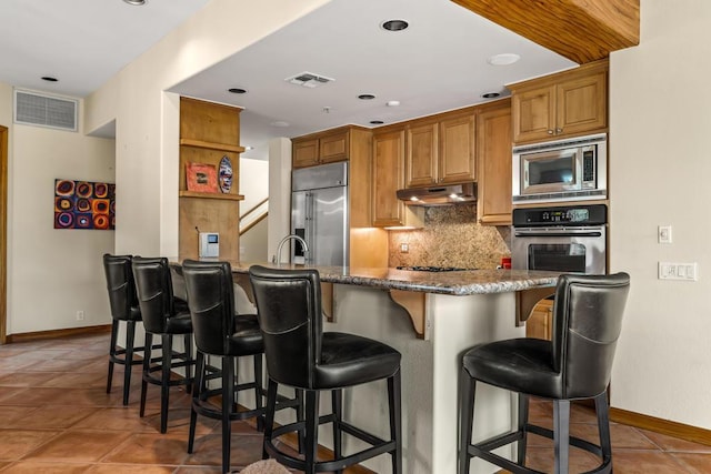 kitchen with a kitchen breakfast bar, tasteful backsplash, dark tile patterned floors, built in appliances, and dark stone counters