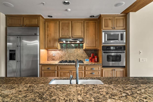 kitchen featuring built in appliances, backsplash, dark stone countertops, and sink