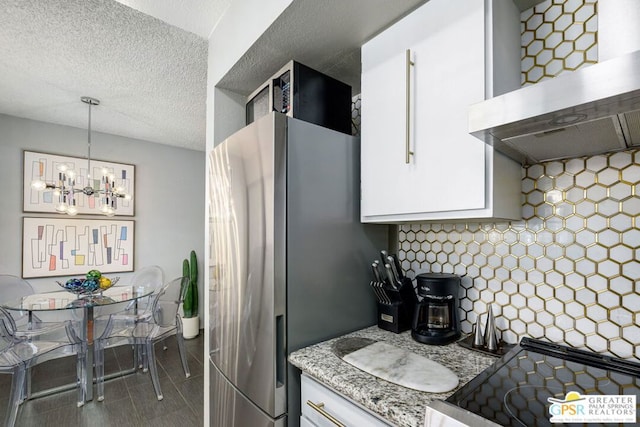 kitchen with a notable chandelier, white cabinets, stainless steel fridge, range hood, and decorative light fixtures