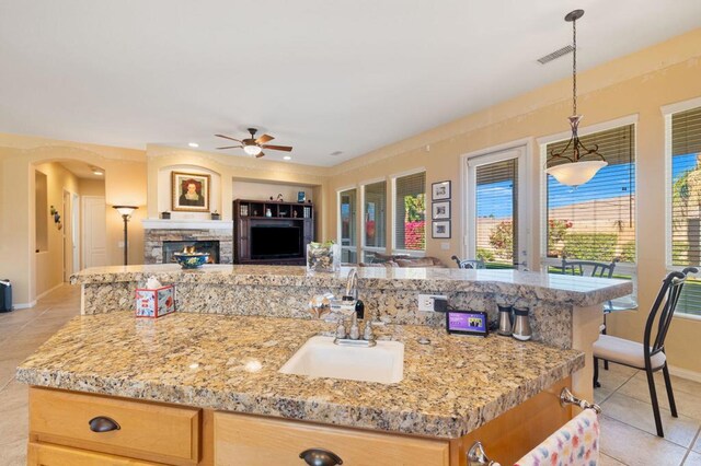 kitchen featuring ceiling fan, sink, hanging light fixtures, an island with sink, and a stone fireplace