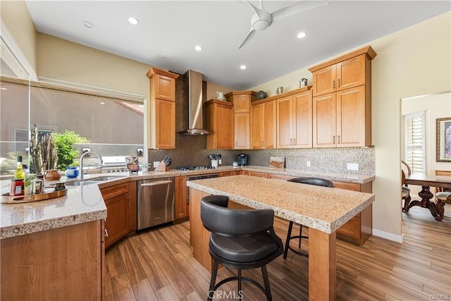 kitchen featuring wall chimney exhaust hood, appliances with stainless steel finishes, light stone countertops, a kitchen bar, and a sink