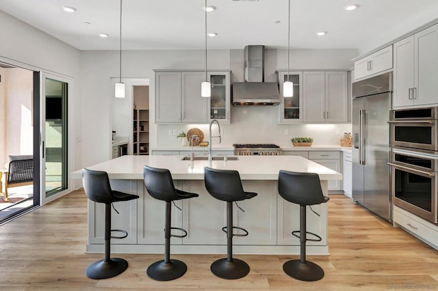 kitchen featuring hanging light fixtures, an island with sink, wall chimney range hood, appliances with stainless steel finishes, and sink