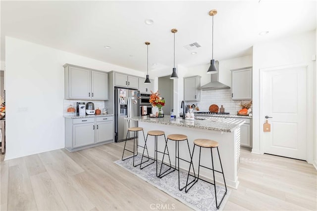 kitchen with decorative backsplash, light stone counters, stainless steel appliances, wall chimney range hood, and a center island with sink