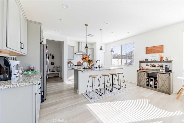 kitchen with gray cabinetry, wall chimney range hood, light stone counters, decorative light fixtures, and a kitchen island with sink