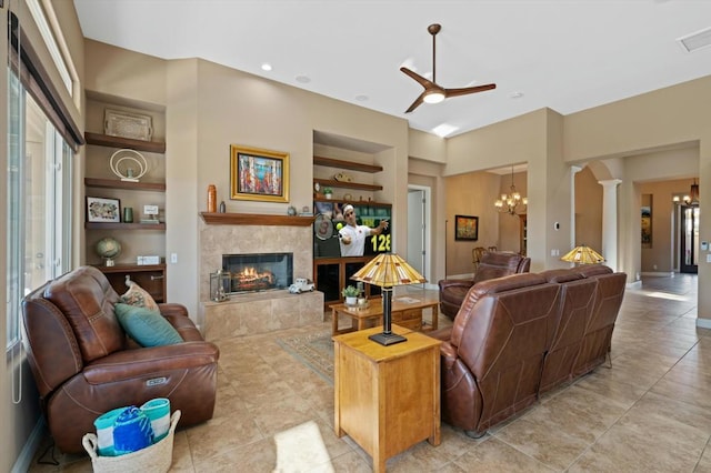 living room with ceiling fan with notable chandelier, built in shelves, a premium fireplace, light tile patterned floors, and ornate columns