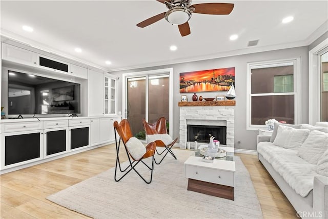 living room with ceiling fan, a stone fireplace, crown molding, and light hardwood / wood-style flooring