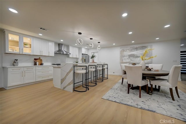 kitchen with glass insert cabinets, hanging light fixtures, light countertops, wall chimney range hood, and white cabinetry