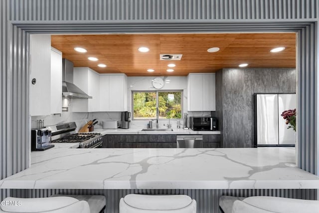 kitchen featuring white cabinets, a kitchen bar, stainless steel appliances, light stone countertops, and wall chimney range hood