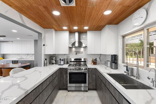 kitchen with sink, white cabinetry, light stone counters, stainless steel range with gas cooktop, and wall chimney range hood