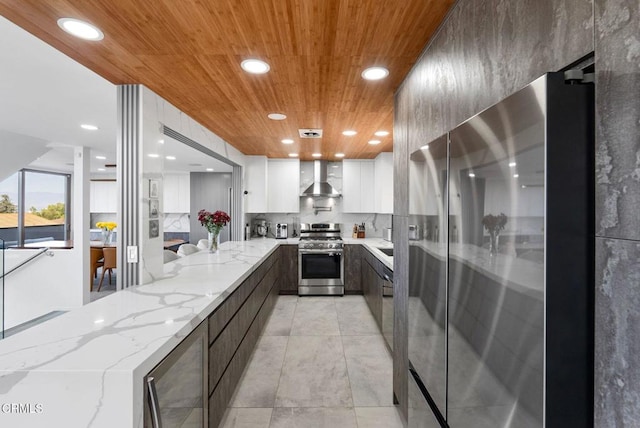 kitchen featuring light stone counters, white cabinets, stainless steel appliances, beverage cooler, and wall chimney range hood