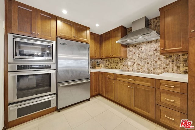 kitchen featuring light tile patterned floors, built in appliances, backsplash, and wall chimney range hood