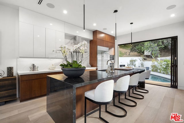 kitchen featuring stainless steel built in refrigerator, white cabinets, an island with sink, and decorative light fixtures