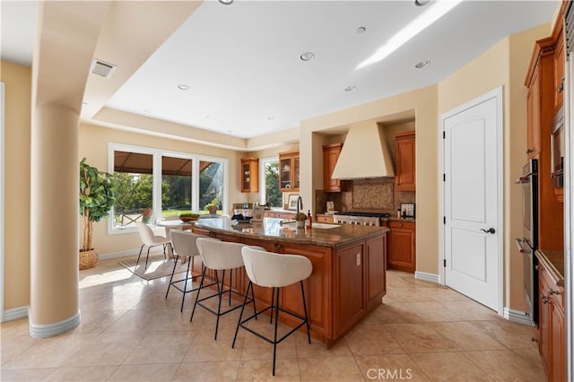 kitchen with light tile patterned floors, premium range hood, dark stone counters, and an island with sink