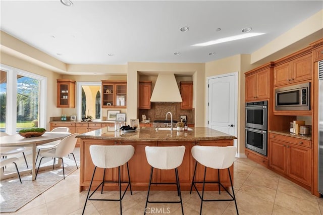 kitchen with premium range hood, stainless steel appliances, a kitchen island with sink, sink, and light tile patterned flooring