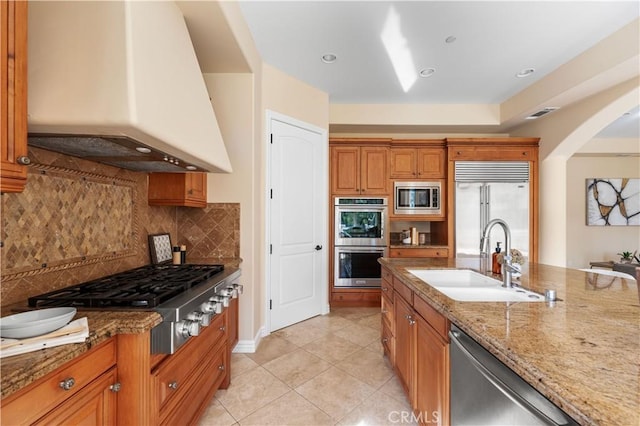 kitchen with light stone countertops, backsplash, premium range hood, sink, and built in appliances