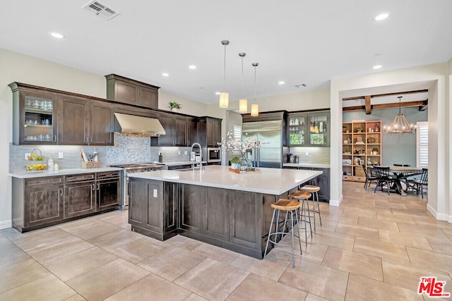 kitchen with beam ceiling, wall chimney range hood, decorative light fixtures, a kitchen island with sink, and dark brown cabinets