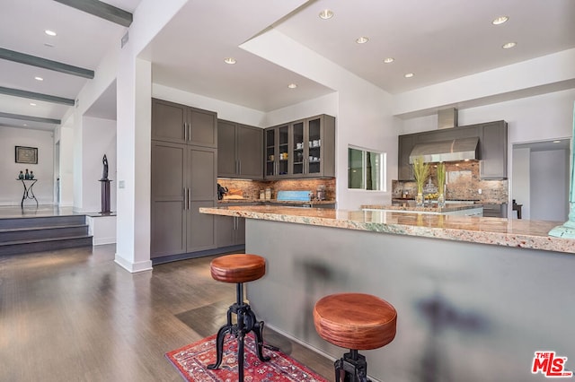 kitchen featuring wall chimney exhaust hood, light stone counters, beamed ceiling, a kitchen bar, and kitchen peninsula