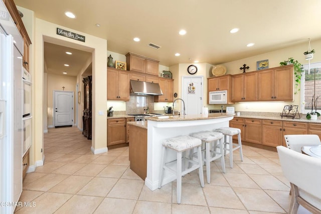 kitchen with a breakfast bar, white microwave, a center island with sink, light stone countertops, and light tile patterned floors