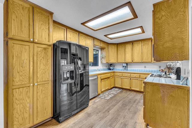 kitchen with tile countertops, black refrigerator with ice dispenser, stainless steel dishwasher, and light hardwood / wood-style flooring