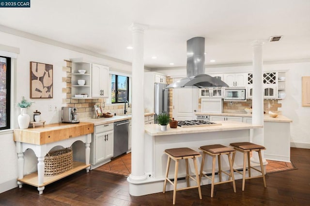 kitchen featuring stainless steel appliances, island range hood, white cabinets, and decorative columns