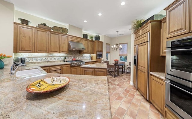 kitchen with light stone countertops, sink, built in appliances, an inviting chandelier, and a kitchen island