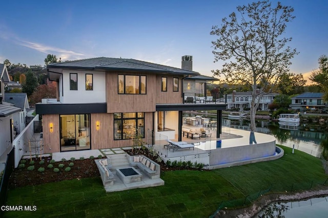 back house at dusk with a lawn, a patio area, a balcony, and a water view