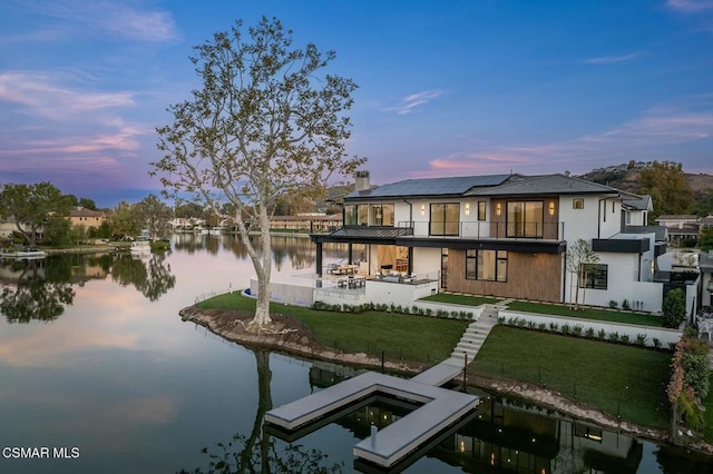 back house at dusk featuring a lawn, a patio area, a water view, and a balcony