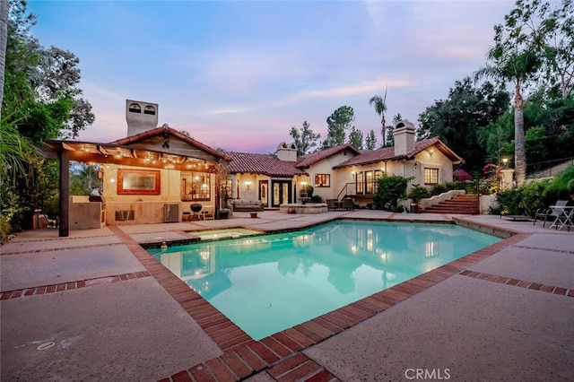 pool at dusk with french doors, a patio, a bar, and an outdoor kitchen