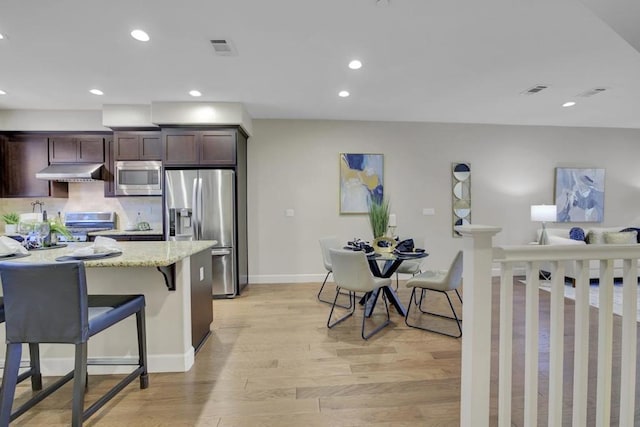 kitchen with light stone countertops, light wood-type flooring, appliances with stainless steel finishes, dark brown cabinetry, and a breakfast bar area