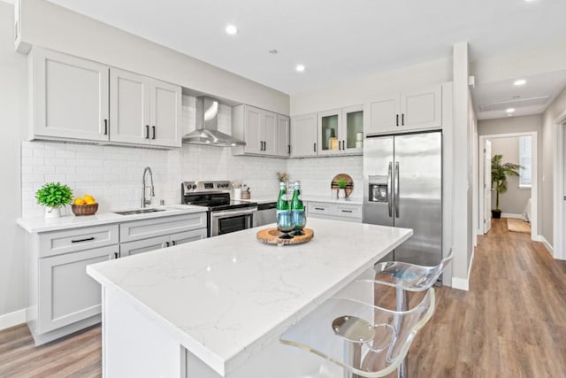 kitchen featuring light stone countertops, wall chimney range hood, decorative backsplash, a breakfast bar, and appliances with stainless steel finishes