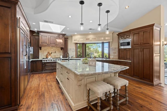 kitchen featuring backsplash, hanging light fixtures, light stone countertops, an island with sink, and appliances with stainless steel finishes