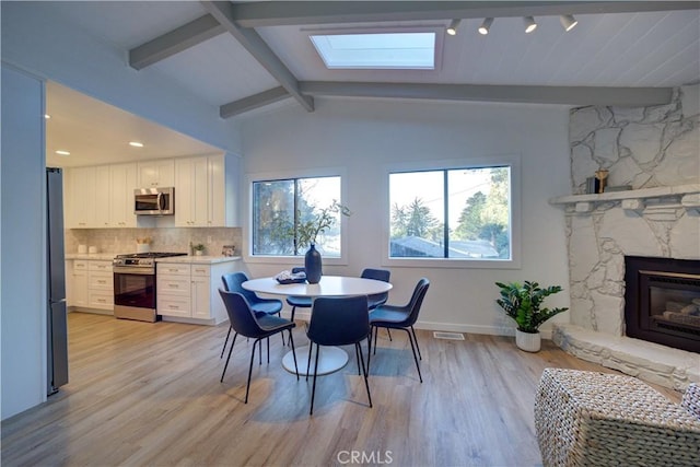 dining space with vaulted ceiling with beams, visible vents, light wood-style floors, a stone fireplace, and baseboards