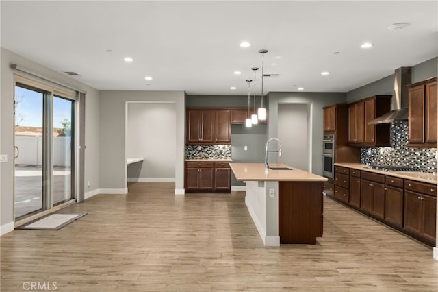 kitchen featuring pendant lighting, a kitchen island with sink, wall chimney range hood, light wood-type flooring, and stainless steel appliances