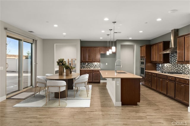 kitchen with stainless steel appliances, hanging light fixtures, wall chimney range hood, an island with sink, and light hardwood / wood-style floors