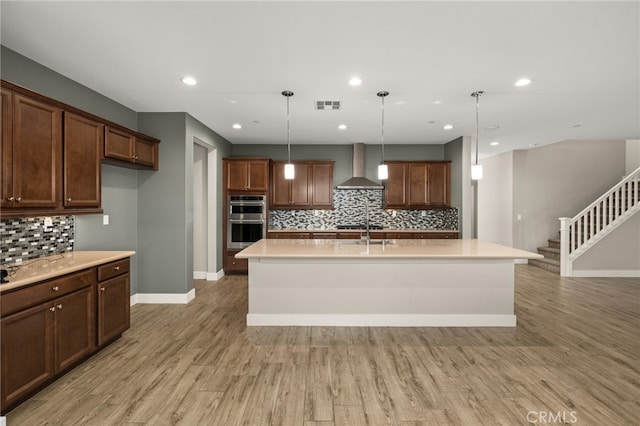 kitchen with double oven, a kitchen island with sink, wall chimney exhaust hood, and decorative light fixtures