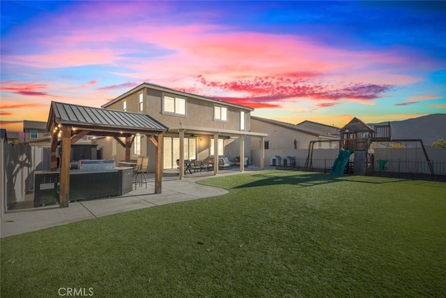 back house at dusk featuring a lawn, a playground, and a patio