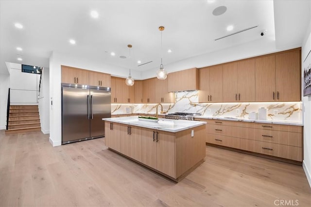 kitchen with tasteful backsplash, an island with sink, pendant lighting, light hardwood / wood-style floors, and stainless steel built in fridge