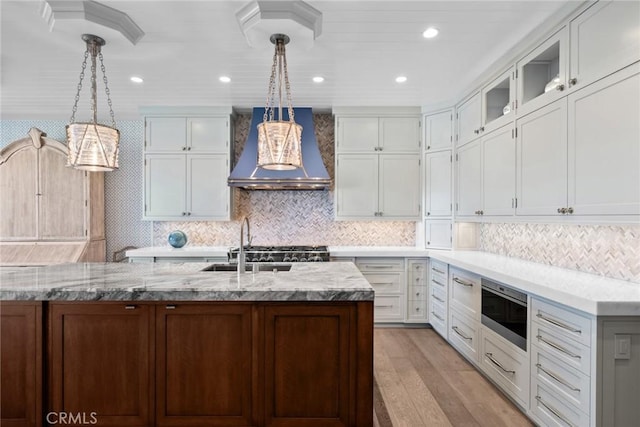 kitchen featuring hanging light fixtures, wall chimney exhaust hood, an island with sink, light stone counters, and white cabinetry