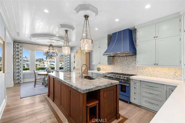 kitchen featuring sink, wall chimney exhaust hood, decorative light fixtures, range with two ovens, and a kitchen island with sink