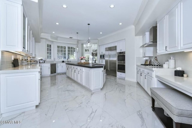 kitchen with built in appliances, hanging light fixtures, wall chimney range hood, a kitchen island, and white cabinetry