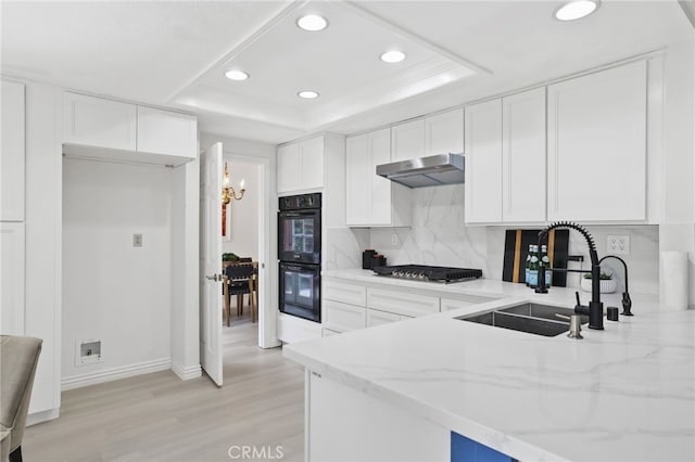 kitchen with a tray ceiling, dobule oven black, stainless steel gas stovetop, a sink, and under cabinet range hood