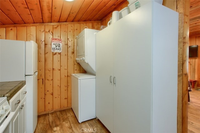 clothes washing area featuring wood walls, stacked washer and dryer, light hardwood / wood-style floors, and wooden ceiling