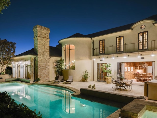 back of house at night featuring a balcony, a patio area, an outdoor pool, and stucco siding