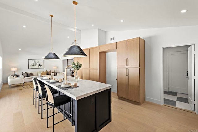 kitchen with light stone countertops, hanging light fixtures, vaulted ceiling, a kitchen island with sink, and light wood-type flooring